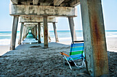 Ein blau, grün und weiß gestreifter Strandstuhl, der leer unter einem Pier am Jupiter Beach, Florida, Vereinigte Staaten von Amerika, steht