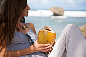 Nahaufnahme einer Frau, die am Strand sitzt und auf den Ozean schaut, während sie eine Kokosnuss mit einem Strohhalm hält,Bathsheba,Barbados,Karibik