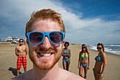 Gruppe von Freunden in Badekleidung am Virginia Beach, mit einem jungen Mann im Vordergrund, der lächelt und eine blau gerahmte Sonnenbrille trägt, First Landing State Park, Virginia, USA, Virginia Beach, Virginia, Vereinigte Staaten von Amerika