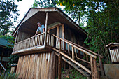 Mann steht auf der Veranda seiner Hütte und blickt in die Kamera, in der Nähe des Bwindi Impenetrable Forest National Park in Uganda, Uganda