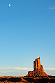 Felsformationen im Monument Valley, Arizona, mit dem Mond vor einem klaren blauen Himmel.  Die roten Felsen leuchten bei Sonnenuntergang, wenn das Licht auf sie trifft, Arizona, Vereinigte Staaten von Amerika
