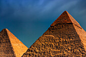 Zwei der Hauptpyramiden von Gizeh, von der Sonne mit einem goldenen Farbton beleuchtet, die Große Pyramide (Cheops) unter einem dunklen, bewölkten Himmel, Gizeh, Ägypten