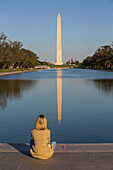 Tourist sitzt am Rande des spiegelnden Beckens mit Blick auf das Washington Monument, aufgenommen vom Lincoln Monument, Washington D.C., Vereinigte Staaten von Amerika