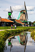 Historische hölzerne Windmühlen, die sich in einer kleinen Gracht mit Fahrradweg und Radfahrern spiegeln,Zaandam,Niederlande