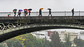 Pedestrians crossing a bridge holding umbrellas on a rainy day,Seville,Spain