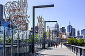 Skyline of buildings in Melbourne viewed from Sandridge Bridge crossing the Yarra River,with sculptures lining the bridge and pedestrians walking towards the waterfront,Melbourne,Victoria,Australia