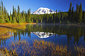 Snow-covered Mount Rainier at sunrise,with autumn coloured vegetation on the shore and mist rising up from the water of Reflection Lake,Washington,United States of America