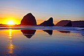 Rock formations along the beach at sunset at Cape Sebastian State Scenic Corridor. A bright sun sinks below the horizon and mirror image reflections are seen in the wet sand at low tide,Oregon,United States of America