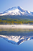 Mirror image of Mount Hood and forest reflected in Trillium Lake with mist rising off the water and people boating and fishing in the tranquil water,Oregon,United States of America