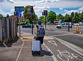 Frau, die einen Koffer auf einem Bürgersteig mit Blick auf den Verkehr in einem Kreisverkehr zieht,Oxford,England