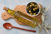Draufsicht auf eine Schale mit Oliven und eine Flasche Olivenöl auf einem Schneidebrett mit Holzlöffel