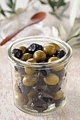 Nahaufnahme eines Glases mit schwarzen und grünen Oliven mit Holzlöffel im Hintergrund