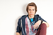 Portrait of teenage boy sitting and leaning against wall,with headphones around neck and looking at camera,studio shot on white background