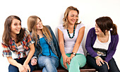 Vier junge Frauen sitzen zusammen auf einer Bank, lachen und schauen sich an, Studioaufnahme auf weißem Hintergrund