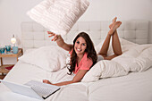 Junge Frau mit Laptop im Bett wirft Kissen in Richtung Kamera
