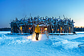 Person steht auf der Brücke, die das Hauptgebäude des beleuchteten Arctic Bath Hotels aus Holzstämmen verbindet, in der Abenddämmerung, Harads, Schwedisch-Lappland, Norrbotten, Schweden, Skandinavien, Europa