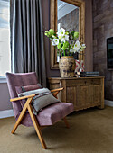 Designer armchair in shades of pink in front of antique wooden chest with ceramic vase and flower arrangement