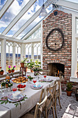 Laid table in a light-flooded conservatory with brick fireplace and plants on windowsill