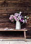 Lilac (Syringa vulgaris) in a metal jug on a wooden bench in front of a wooden wall