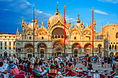 Die Basilika San Marco spiegelt sich im Wasser auf der Piazza San Marco in der Dämmerung während des Sonnenuntergangs, Venedig, Italien, mit Bewegungsunschärfe auf den Touristenmassen