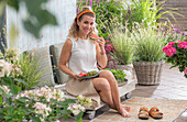 Woman eating melons on the terrace between flower pots with grass lily 'Starlight' (Anthericum), hydrangea (Hydrangea), purple loosestrife (Lythrum salicaria)