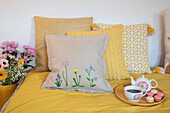 Embroidered cushion with spring flowers on yellow bedspread, tea set and bouquet of flowers