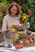 Woman decorates Thanksgiving cake stand made of basket and birch slices with zucchini, apples, eggplant, marigold, tomato, wax bean, thyme, sage; rosemary, walnut; Mini pumpkin, peppers on a set table