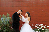 Bräutigam küsst die Hand der Braut vor einer Mauer und Blumen