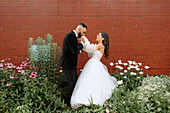 Bräutigam küsst die Hand der Braut vor einer Mauer und Blumen