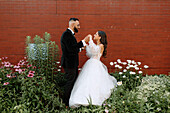 Bride and groom holding hands against brick wall and flowers