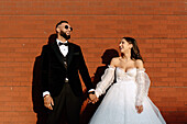 Smiling bride and groom holding hands against brick wall