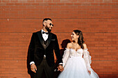 Smiling bride and groom holding hands against brick wall