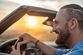 Lächelnder Mann am Steuer eines Cabriolets in einer Landschaft bei Sonnenuntergang