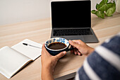 Nahaufnahme der Hände einer Frau, die eine Kaffeetasse am Schreibtisch mit Laptop und Notizblock hält
