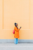 Italien, Mailand, Frau mit Kopfhörern und Smartphone vor orangefarbener Wand