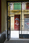 Ein offenes Metalltor im unteren Stockwerk eines Wohnhauses mit Graffiti an der Tür gegenüber; Barcelona, Spanien
