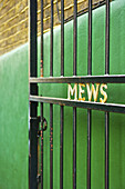 Ein offenes Metalltor mit dem Wort Mews" in goldener Schrift vor einer grünen Wand; London, England".