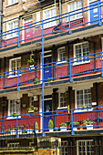Ein Wohngebäude aus Backstein mit blauem Balkongeländer und Türen sowie einem roten Streifen auf der unteren Hälfte jeder Wand; London, England.