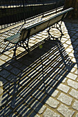 A Bench And Railing With Shadows Cast On A Walkway; Paris, France