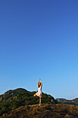 A Young Woman Does A Yoga Pose On A Hilltop Against A Blue Sky; Ulpotha, Embogama, Sri Lanka