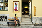 A Woman Sits On A Bench Outside A Yellow Building; Athens, Greece