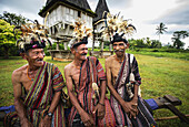 Gruppe von Männern in traditioneller Kleidung lacht gemeinsam mit heiligen Häusern im Hintergrund; Bezirk Lospalmos, Timor-Leste