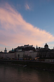 Das historische Zentrum und das Schloss von der Salzach aus gesehen in der Abenddämmerung mit einer rosa Wolke; Salzburg, Österreich