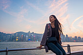 A Young Woman At The Waterfront Sitting On The Railing At Sunset With The Skyline In The Background, Kowloon; Hong Kong, China