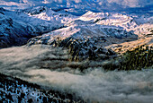 Morning fog fills the valley between snowy, white peaks of the stark and rugged San Juan Mountains. Shafts of silver, not sunlight, lured miners into Colorado's wilderness where now rugged trails form the Alpine Loop, a Bureau of Land Management back country byway with more than a glimmer of mountain splendour; Colorado, United States of America
