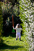 Rückenansicht eines jungen Mädchens, das in einem Garten spazieren geht und eine Spielzeugharke hält