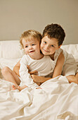 Junge und Kleinkind sitzen im Bett, umarmen sich und lächeln