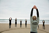 Back view of a surfing instructor and a group of students stretching their arms on a beach