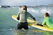 Rückenansicht einer Frau im Meer, die ein Surfbrett hält, auf dem ein Mädchen liegt