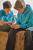 Two teenage boys sitting on a bale of hay sharing an mp3 player